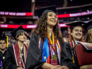 A smiling Stephanie Viera '25 is seated among graduates at the 2025 Commencement wearing a graduation robe, and several stoles, cords, and medals