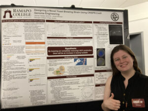 Student posing in front of their research poster on designing a novel yeast brewing strain using CRISPR-Cas9 genome engineering, SCHOLARS' DAY, April 16th, 2024.